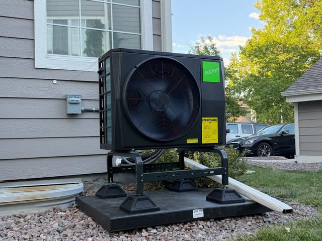 A 2-ton Jetson Air ducted heat pump on a black metal riser stand along the gray vinyl-sided exterior of a home in Westminster, CO, with a dedicated disconnect box mounted to the wall beside it.