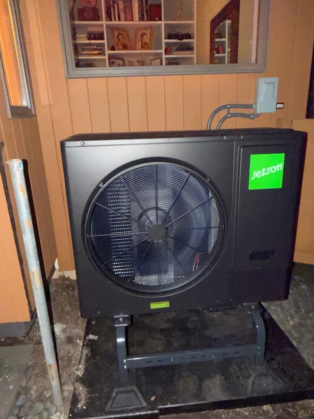Jetson Air 3-ton central ducted heat pump installed on a black metal stand against tan vertical siding on a home in White Plains, NY, with conduit running up to a wall-mounted disconnect box.