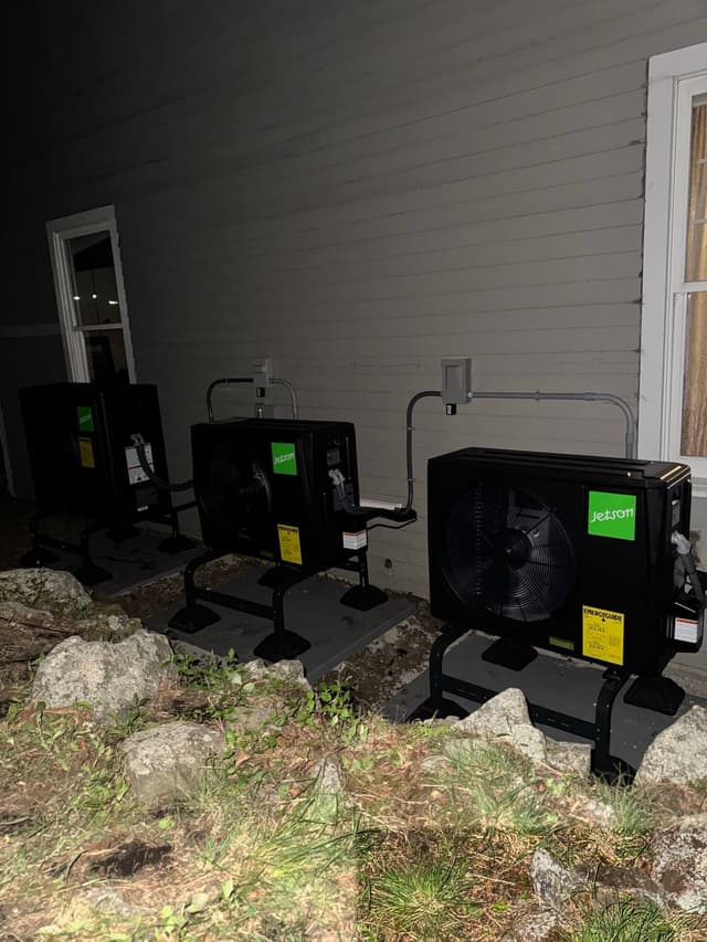 Three black Jetson Air heat pump condensers mounted in a row along the side of a light-gray-sided house in Winchester, MA, photographed at night.