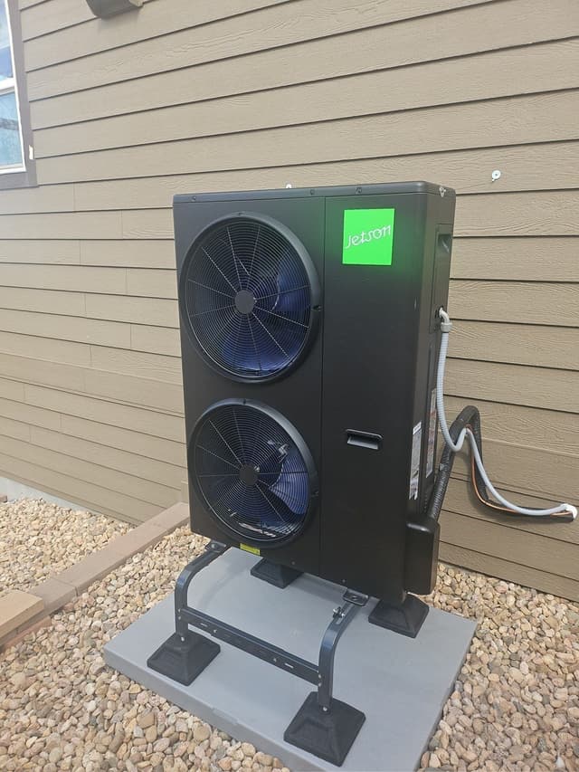 Black Jetson Air 5-ton heat pump condenser on a metal riser stand on a concrete pad with decorative gravel, mounted against tan lap siding in Windsor, CO.