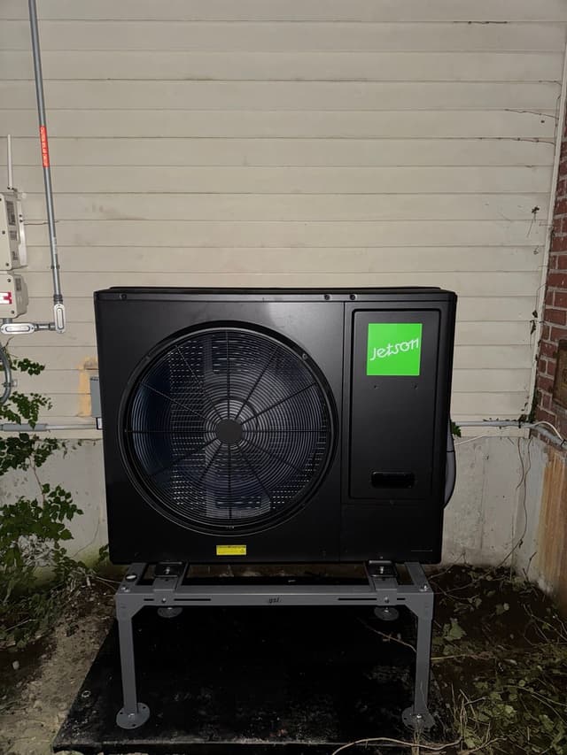 Black Jetson Air 3-ton central ducted heat pump condenser installed along the side of a house with white vinyl siding in Woburn, MA.