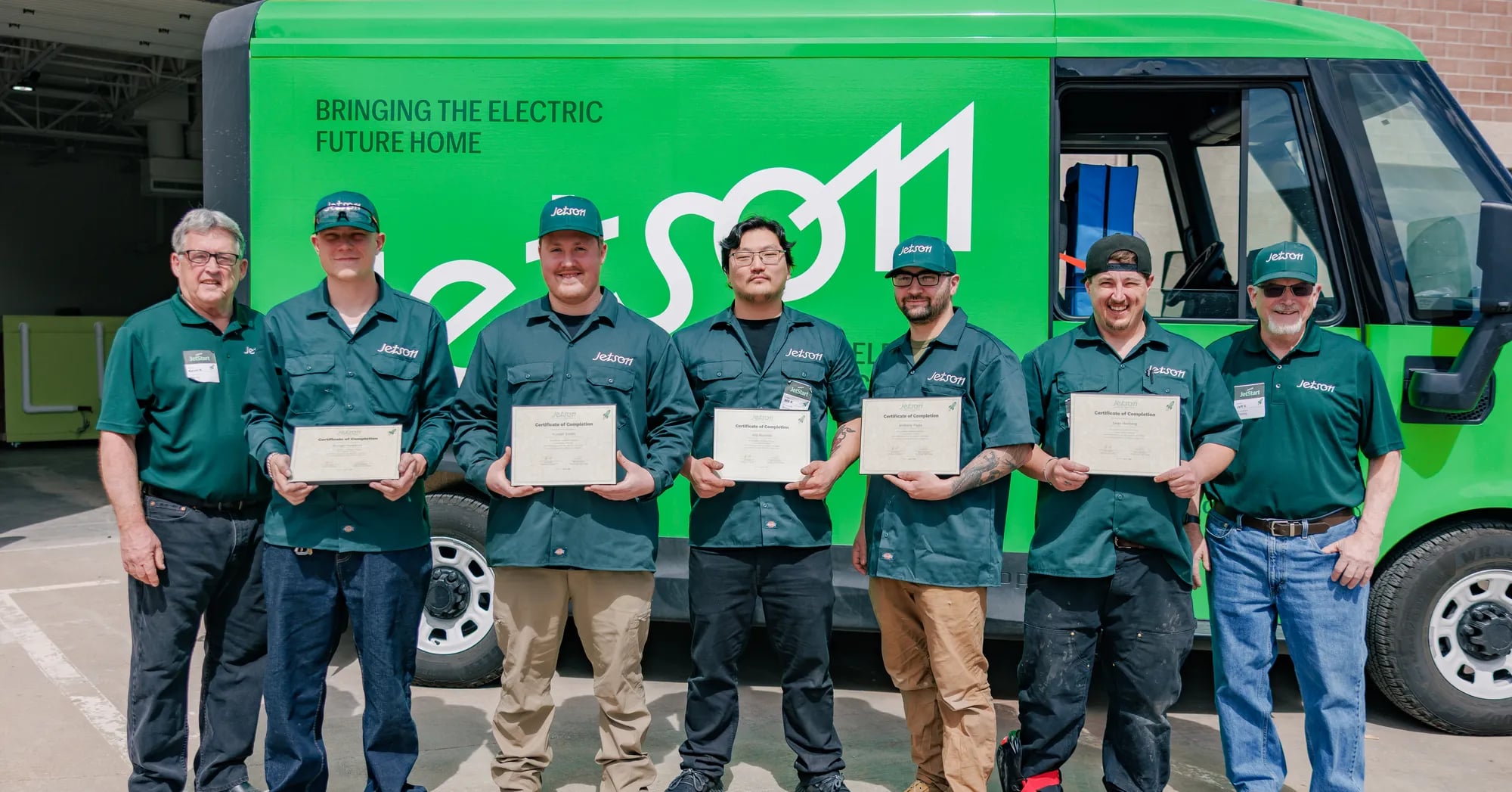 A graduating class of Jetson technicians in uniform holding their certificates of completion in front of a green Jetson van