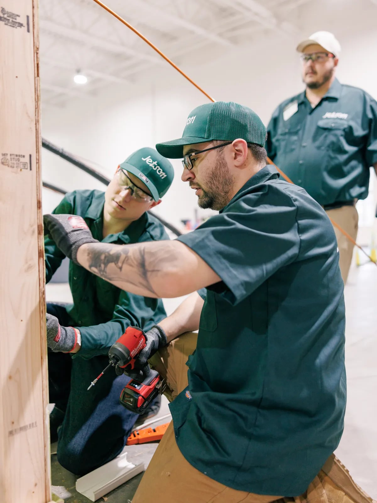 A Jetson trainee drilling a framed wall while an instructor looks on