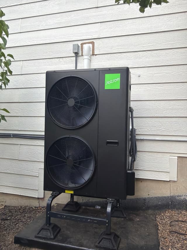 Black Jetson Air 4-ton heat pump condenser mounted against white vinyl siding of a home in Arvada, Colorado, with refrigerant lines and electrical conduit running into the wall.