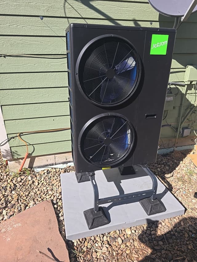 Jetson Air 4-ton dual-fan outdoor heat pump unit mounted on a polymer riser pad on a gravel bed beside a green-sided home in Arvada, CO, with conduit and refrigerant lines neatly finished.