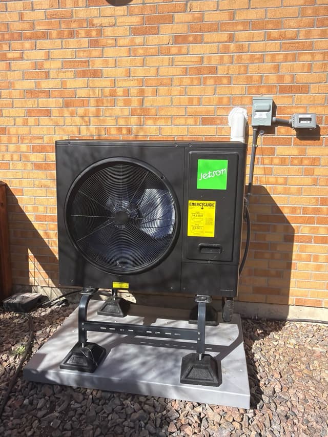 Jetson Air 3-ton central ducted heat pump on a metal stand over a concrete pad with decorative rock landscaping, mounted against a red brick exterior wall in Arvada, CO.