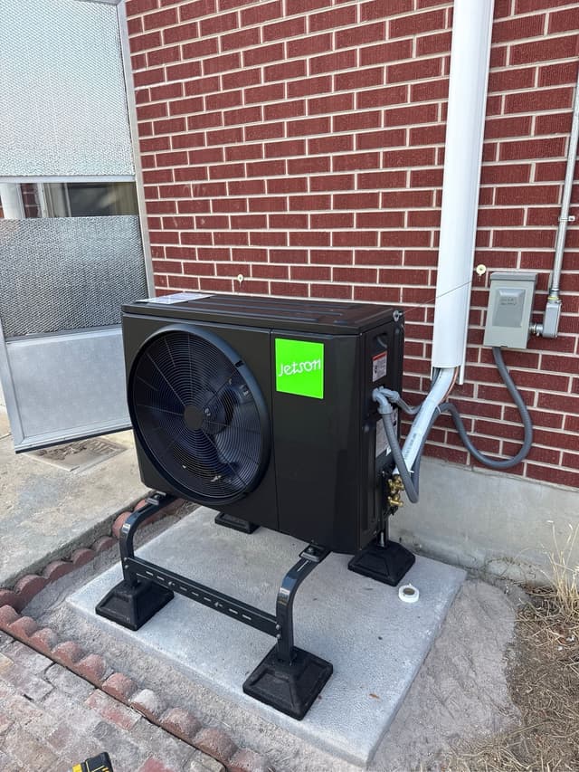 2-ton Jetson Air central ducted heat pump unit installed against a red brick exterior wall in Arvada, Colorado, with refrigerant lines and conduit running up to a wall-mounted disconnect box.