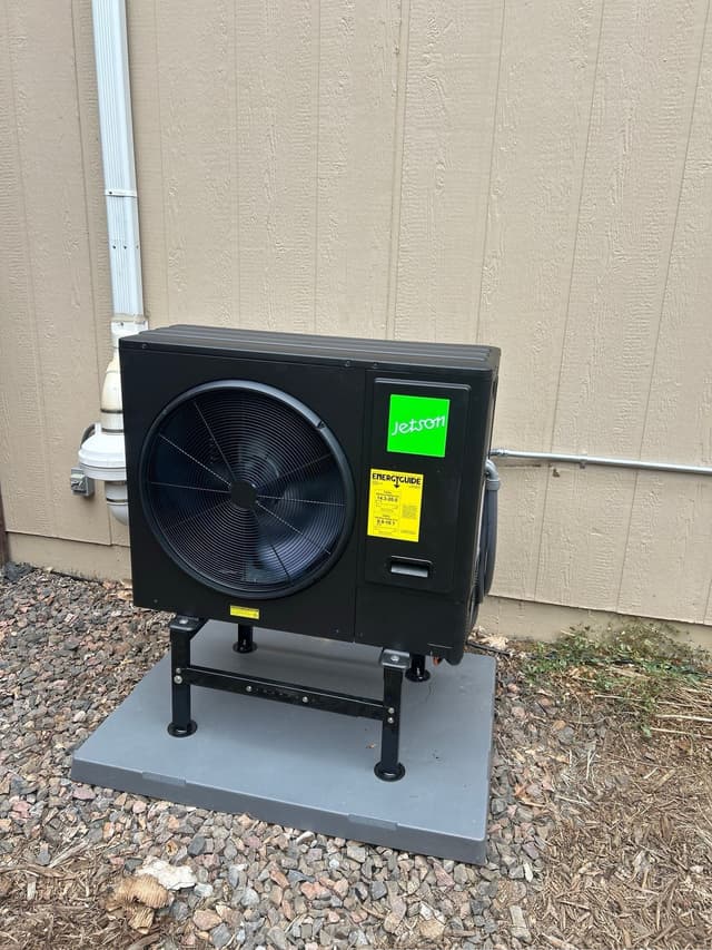 Black Jetson Air 3-ton heat pump condenser installed against a tan board-and-batten building in Arvada, Colorado, with refrigerant lines running along the wall.
