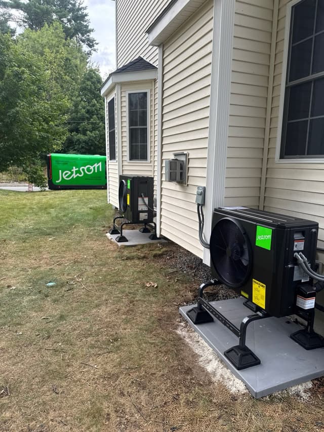 Two black Jetson Air heat pump condensers installed side by side along the exterior of a beige vinyl-sided home in Attleboro, with a green Jetson-branded truck visible in the background.