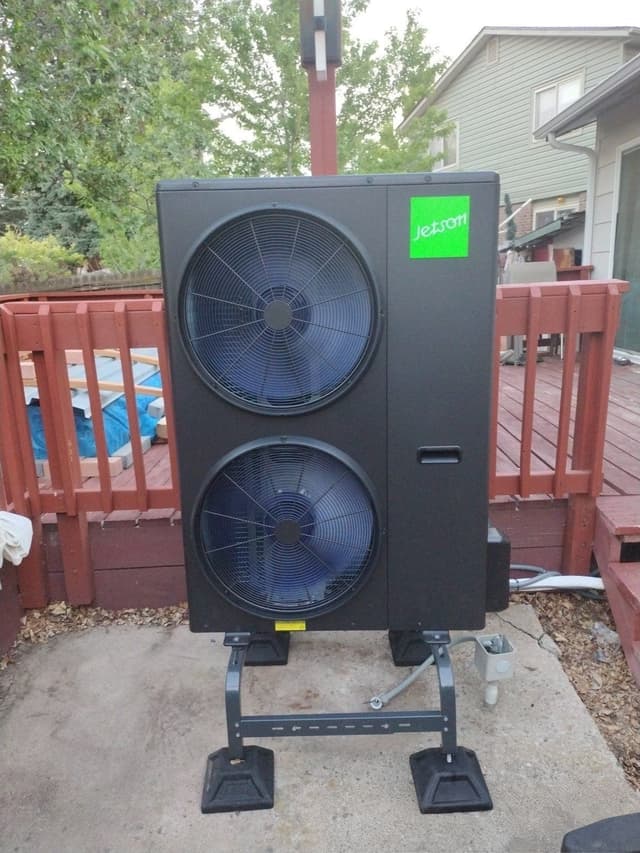 Jetson Air dual-fan heat pump condenser on a black metal stand with rubber pads, mounted on a concrete patio beside a red wood deck railing in Aurora, CO.