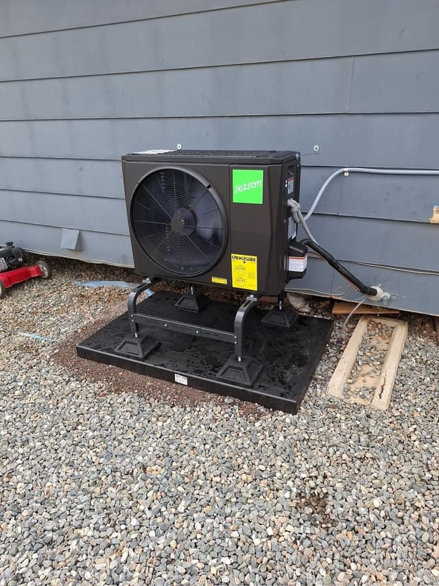 Black 2-ton Jetson Air heat pump condenser installed against gray horizontal siding at an Aurora home, with refrigerant lines running into the wall.