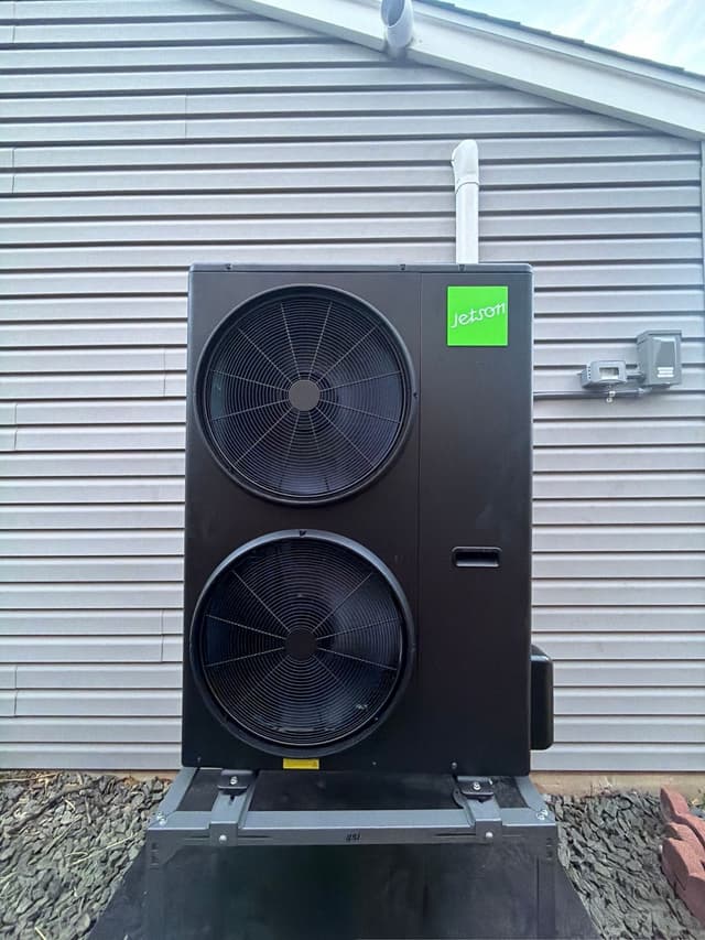 A large black dual-fan Jetson Air heat pump unit installed against grey horizontal-lap siding on an Aurora home, with a white conduit running up the wall and a grey electrical disconnect box mounted nearby.