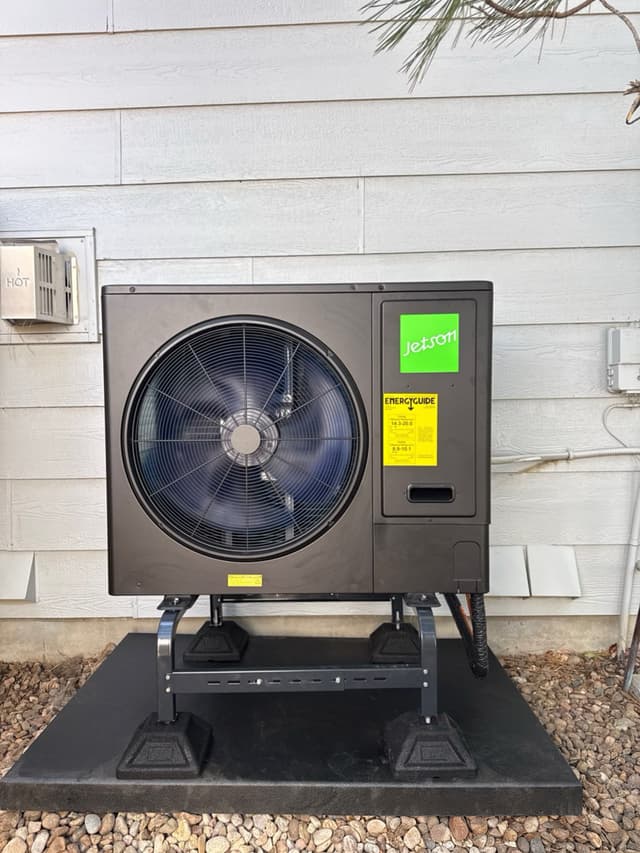 Jetson Air 3-ton heat pump condenser on an elevated metal stand against gray house siding in Aurora, CO, with EnergyGuide sticker and disconnect box visible.
