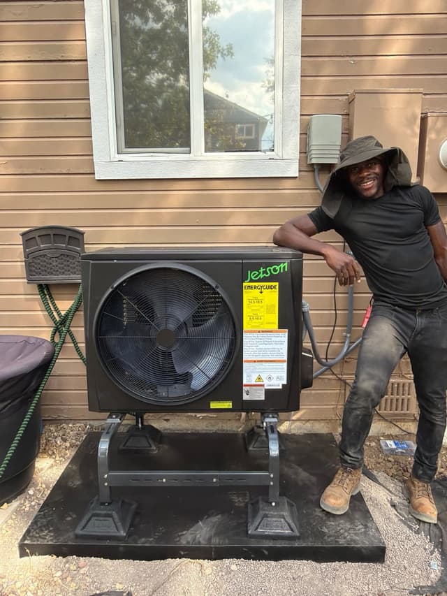 Crew member smiling and leaning against a newly installed Jetson Air 2-ton central ducted heat pump on the exterior of a tan lap-siding house in Aurora, Colorado.