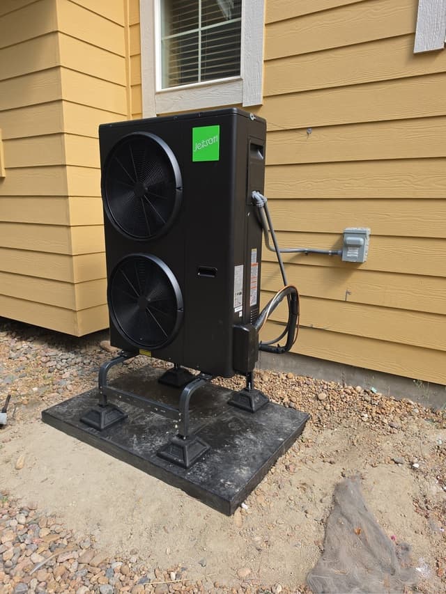 Black Jetson Air dual-fan 4-ton heat pump condenser mounted against yellow lap siding on a home in Aurora, with electrical conduit and refrigerant lines running to a disconnect box on the wall.