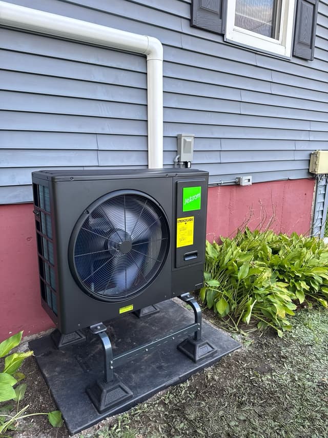 Black 3-ton central ducted heat pump condenser installed against gray lap siding with a red foundation in Billerica, MA, with hosta plants visible to the right.