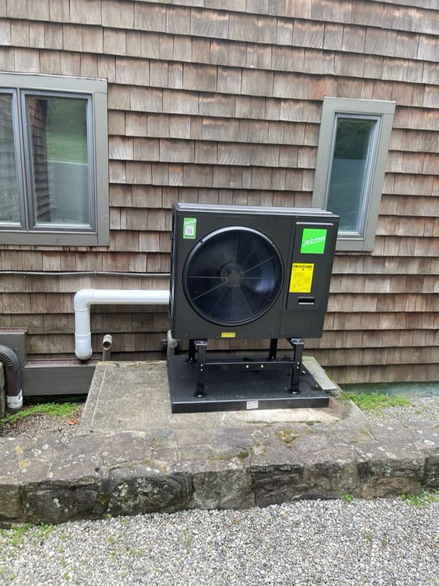 Black Jetson Air 3-ton heat pump condenser on a black metal stand and concrete pad alongside the exterior of a wood-shingle house in Briarcliff Manor, NY.