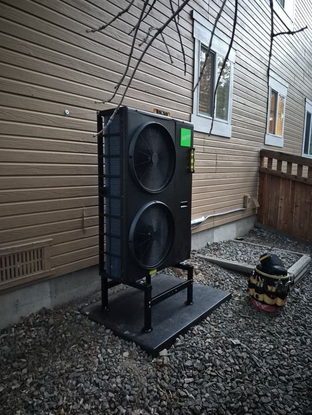 4-ton Jetson Air central ducted heat pump unit installed along the side of a tan house in Brighton, with a tool bag resting on the gravel beside it.