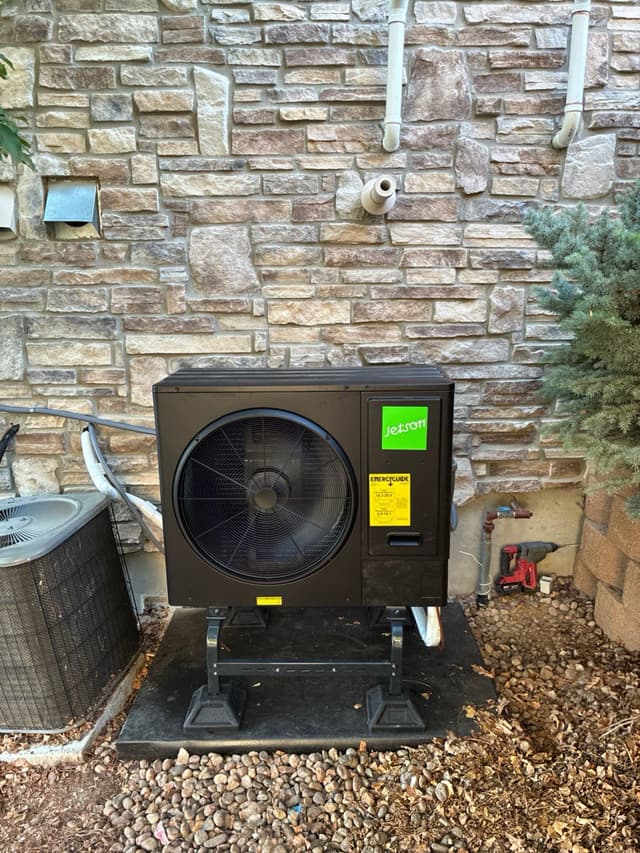 Jetson Air 3-ton heat pump mounted on a metal stand over river rock against a natural stone veneer wall at a Broomfield, CO home, with conduit and PVC piping running up the wall behind it.