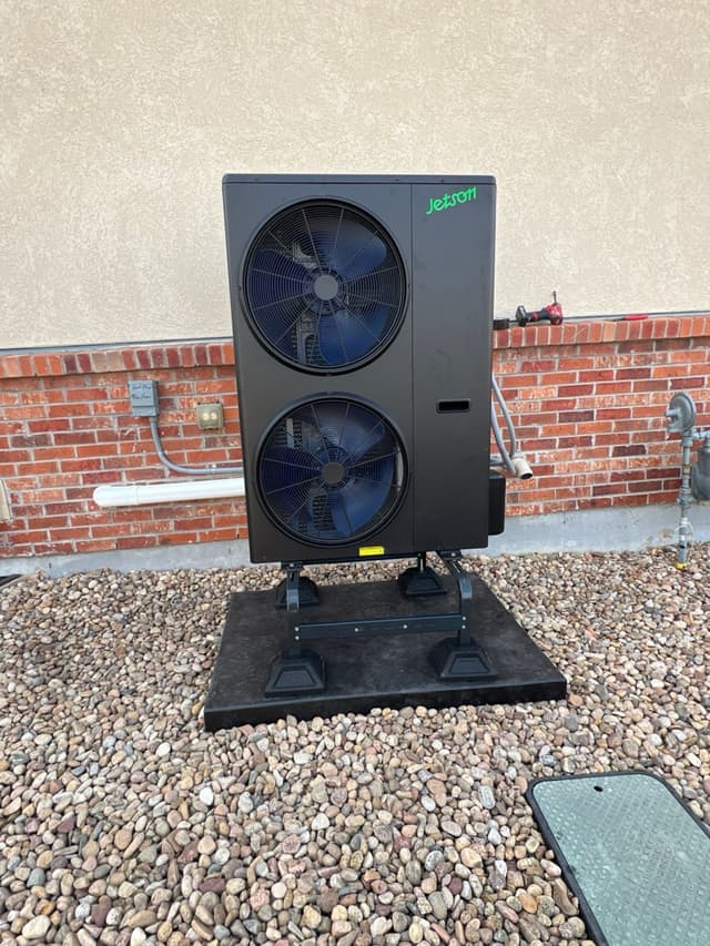 A black Jetson Air dual-fan heat pump condenser mounted on adjustable legs over a river-rock gravel bed against a stucco exterior wall in Broomfield, CO.