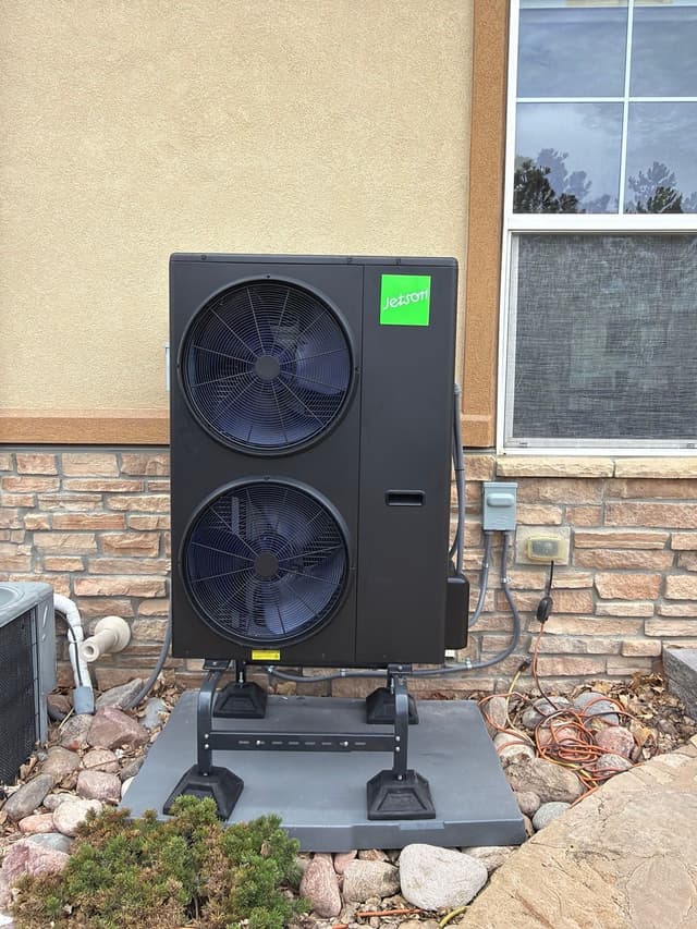 A large black dual-fan heat pump condenser installed against the stucco-and-stone exterior wall of a home in Broomfield, CO, with electrical conduit running to a weatherproof disconnect box on the wall.