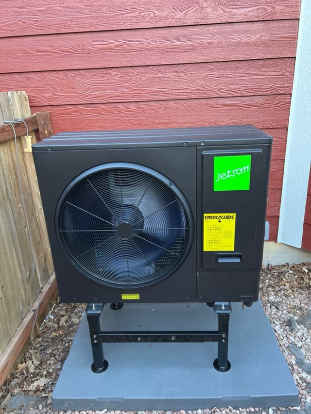 Jetson Air 3-ton central ducted heat pump unit mounted in a gravel bed between a wooden fence and white trim, with red horizontal lap siding in the background, in Broomfield, CO.