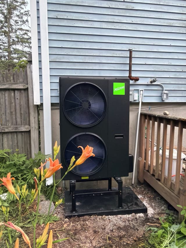 A black Jetson Air dual-fan outdoor heat pump condenser on an elevated metal stand over a gravel pad beside a light-blue house, with orange day lilies blooming in the foreground.