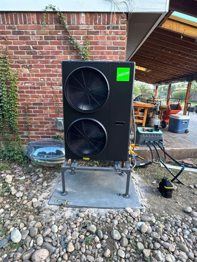 A black Jetson Air dual-fan 4-ton heat pump installed in a backyard patio in Centennial, CO, with a red brick wall covered in ivy in the background.