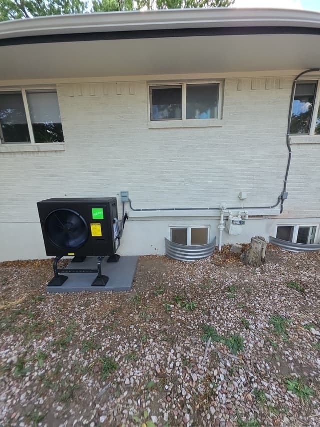 Black Jetson Air 3-ton heat pump condenser mounted on the side of a single-story white brick home in Colorado Springs, with electrical conduit running neatly along the wall.