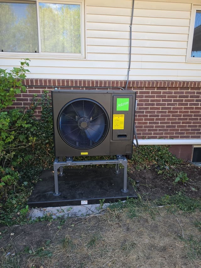 Black Jetson Air 3-ton heat pump condenser installed along the side of a Colorado Springs home with red brick foundation and cream vinyl siding, with refrigerant lines running up the wall.