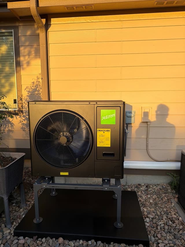 Black Jetson Air 3-ton heat pump condenser installed along the side of a tan lap siding home in Commerce City North, Commerce City, with electrical disconnect and conduit visible on the wall.