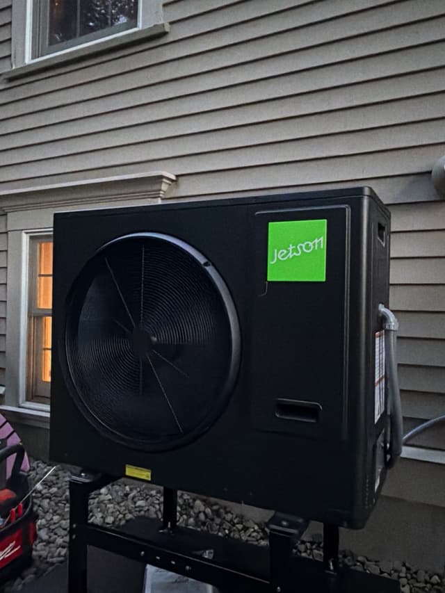 Black Jetson Air 3-ton heat pump condenser on a metal stand against gray vinyl siding, shot at dusk with refrigerant lines exiting the right side.