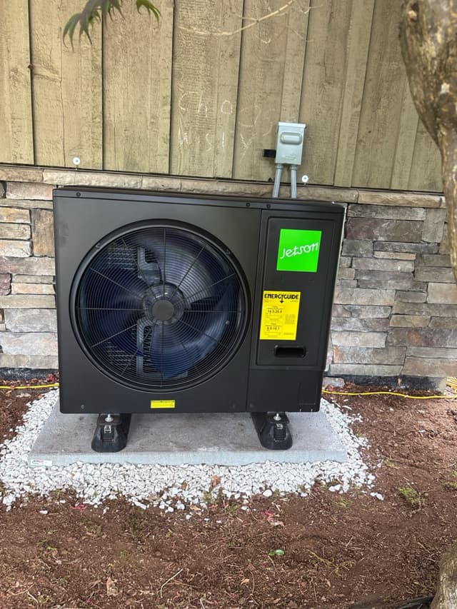 Jetson Air 3-ton central ducted heat pump on a concrete pad with white gravel, mounted against a stone-veneer wall with disconnect box and conduit, wooden fence in background, Tsawwassen, Delta, BC.