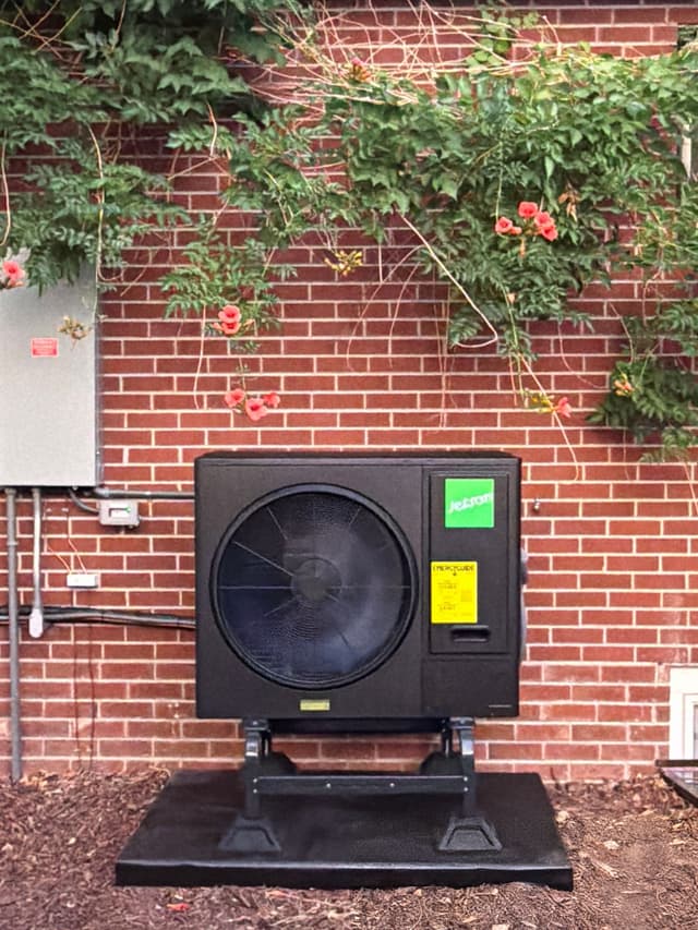 Jetson Air 3-ton central ducted heat pump mounted against a red brick wall with orange-red trumpet vine flowers draped along the top, electrical disconnect and conduit visible to the left, Denver, CO.