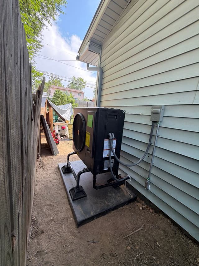 A black Jetson Air 3-ton heat pump condenser installed on a pad in a narrow side yard between a wood fence and light-blue vinyl-sided house in Denver, CO, with conduit and refrigerant lines running up to a wall-mounted disconnect box.