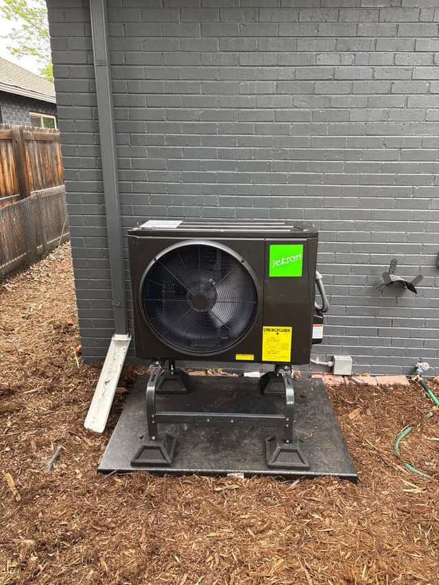 Black Jetson Air outdoor heat pump unit on a stand and pad along the side of a dark gray brick house in Denver, with refrigerant lines and an electrical disconnect visible against the wall.