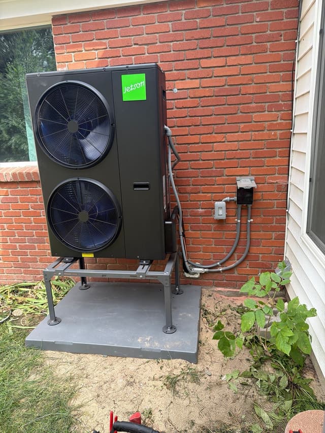 A black Jetson Air 5-ton central ducted heat pump mounted against a red brick wall at a home in Denver, with electrical conduit running to a disconnect box.