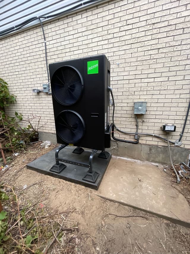 Black Jetson Air 4-ton heat pump installed on a concrete pad against a light brick exterior wall in Denver, with electrical conduit and disconnect box mounted to the wall.