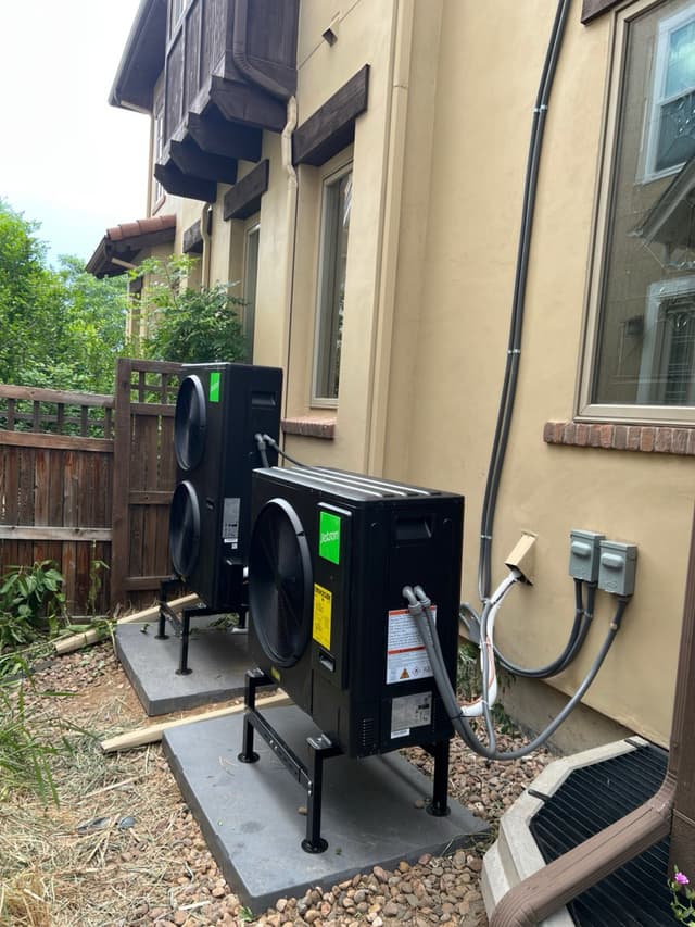 Two black Jetson Air heat pump condensers mounted side by side on metal pad stands along the stucco exterior of a Denver home, with conduit and refrigerant lines running up the wall to disconnect boxes.