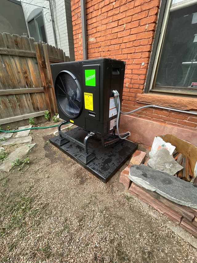 Black Jetson Air 3-ton central ducted heat pump installed in a side yard against a red brick building in Denver, Colorado.