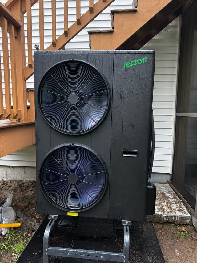 Jetson Air 5-ton heat pump mounted on a metal stand outside a home with white vinyl siding in Dudley, MA, wet from recent rain.