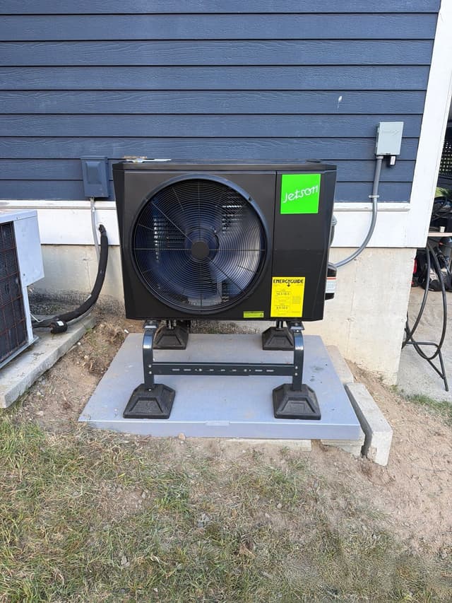 Black Jetson Air outdoor heat pump condenser installed against dark blue horizontal siding on a home in Dunstable, MA, with electrical conduit running up to a disconnect box.