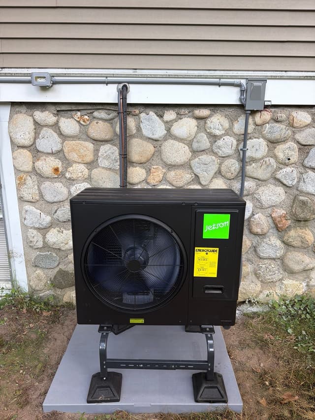 Jetson Air 3-ton outdoor heat pump unit installed against a fieldstone foundation wall with vinyl siding above, refrigerant lines and conduit running neatly to a disconnect box, in Dunstable, MA.