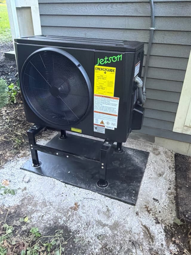 Jetson Air 2-ton outdoor heat pump unit mounted on a black metal riser on a ground-level pad beside a gray-sided house in Easton, MA.