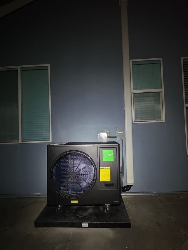 Black Jetson Air 3-ton heat pump condenser installed against a blue-grey exterior wall in Elk Grove, with line-set conduit running up to a disconnect box above the unit.