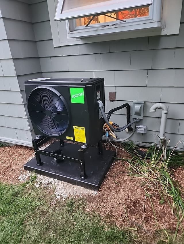 A black Jetson Air 2-ton central ducted heat pump mounted on a metal stand against gray shingle siding in the corner of a home in Elmsford, NY, with line sets and conduit running to an electrical disconnect box.