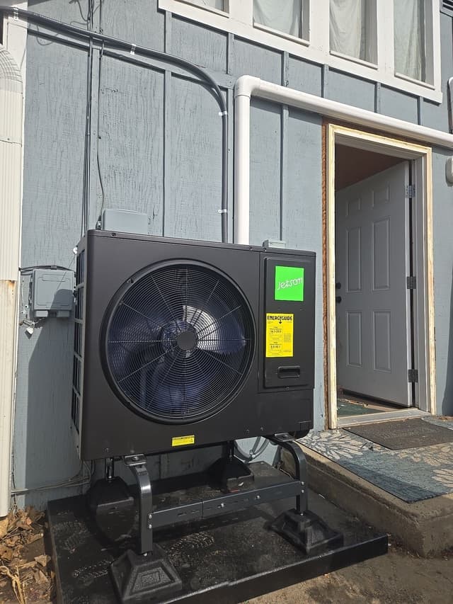 Black Jetson Air 3-ton heat pump condenser on a metal stand and concrete pad beside a light-blue-gray sided house in Erie, CO, with white line-set covers and conduit running neatly up the wall to a disconnect box.