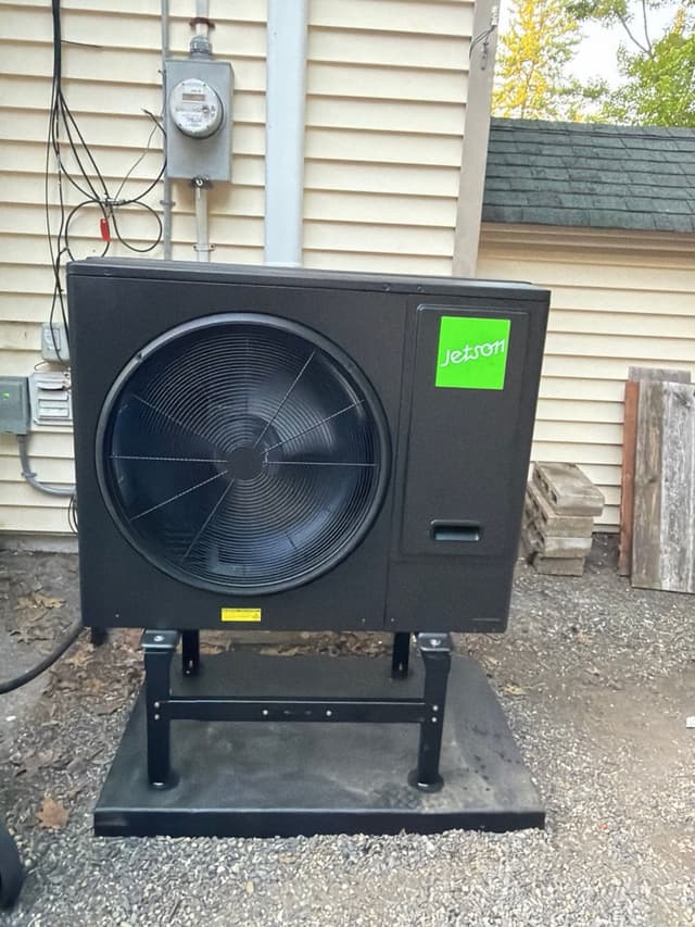 Jetson Air 3-ton central ducted heat pump mounted on an elevated black metal stand along the side of a home with beige vinyl siding in Fitchburg, MA.