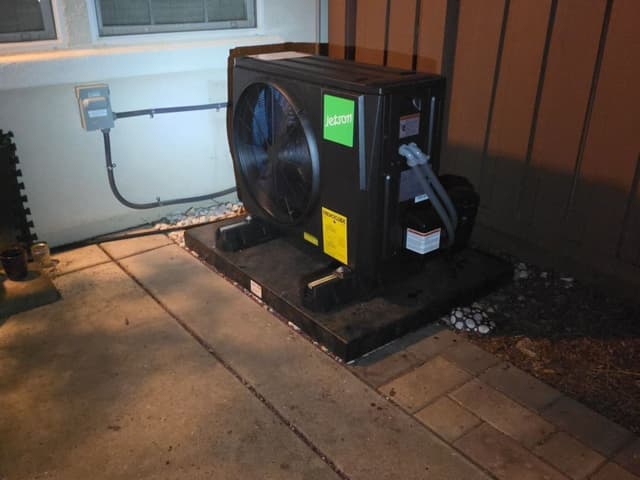Black Jetson Air 2-ton central ducted heat pump installed against a stucco wall with a wooden lattice fence, refrigerant lines exiting the side and an electrical disconnect box mounted nearby, Folsom, CA.