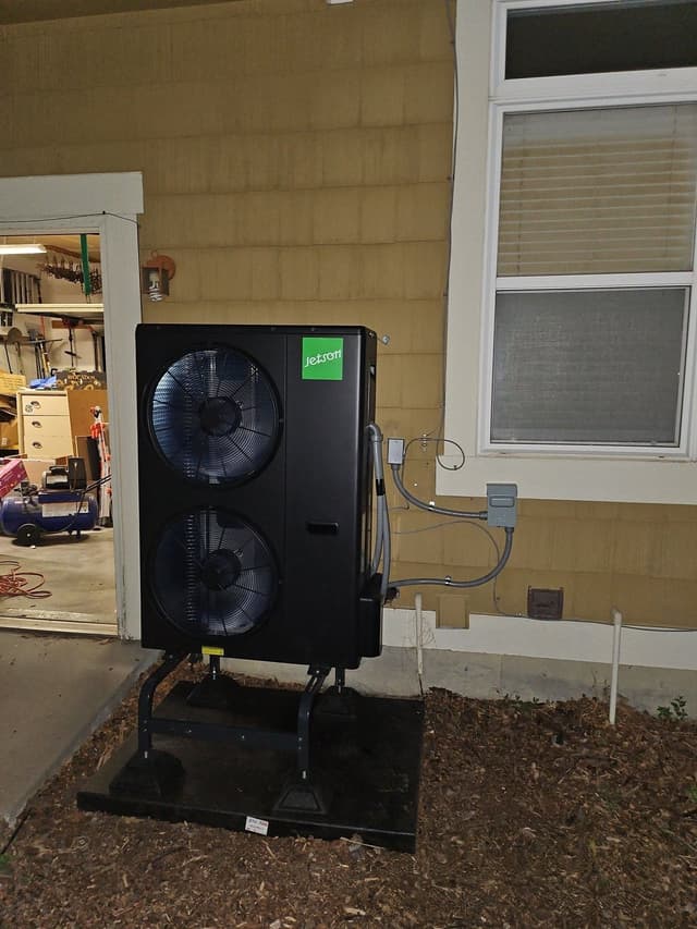 Black dual-fan heat pump condenser on a black equipment pad in a mulched bed beside a house wall in Fort Collins, CO, with electrical conduit running to a disconnect box.